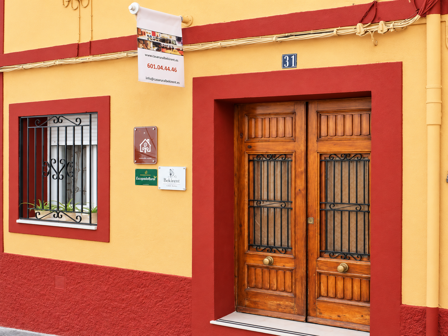 Fachada de Casa Rural Bekirent en Bocairent, Valencia, con puerta doble de madera, ventana con rejas y paredes amarillas con detalles rojos.